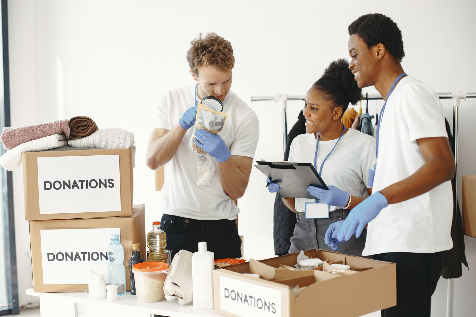 Three diverse volunteers in white shirts and blue gloves sorting cardboard donation boxes filled with food and supplies at a brightly lit indoor table.