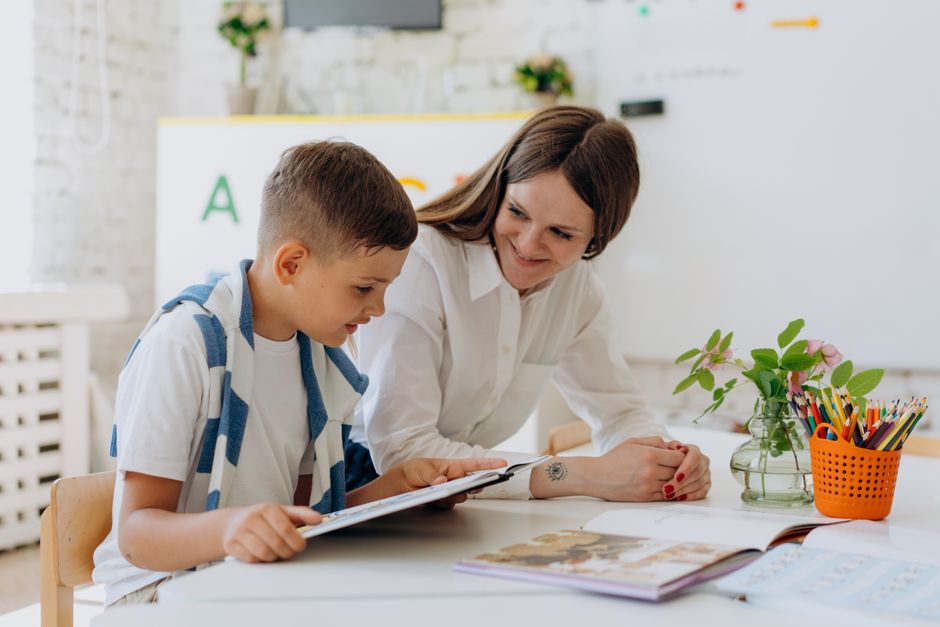 A smiling teacher leaning over a desk helping a young school-age boy with a tablet in a bright classroom, with a flower vase and colored pencils on the desk.