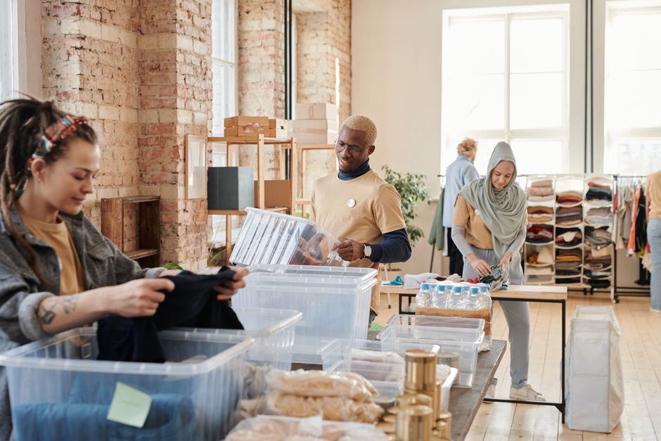 Diverse volunteers sorting donated clothing into clear plastic bins in a bright loft with exposed brick and a clothing rack in the background.