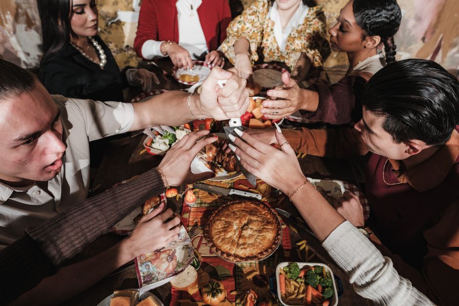 A diverse group gathered around a festive table with a golden pie in the center, hands reaching in to share a community meal together.