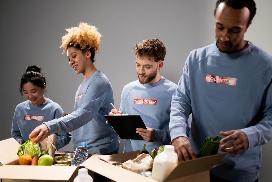Four diverse volunteers in blue volunteer shirts packing a food donation box with fresh produce and smiling.