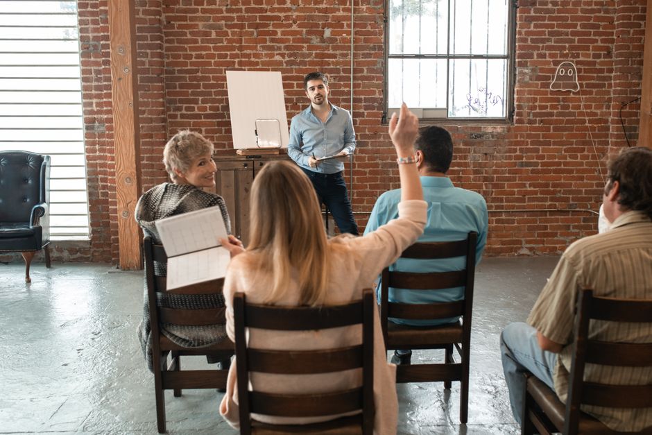 A male instructor stands at a flipchart leading a workshop for four diverse adult participants in a brick-walled room, with one participant raising a hand to speak.