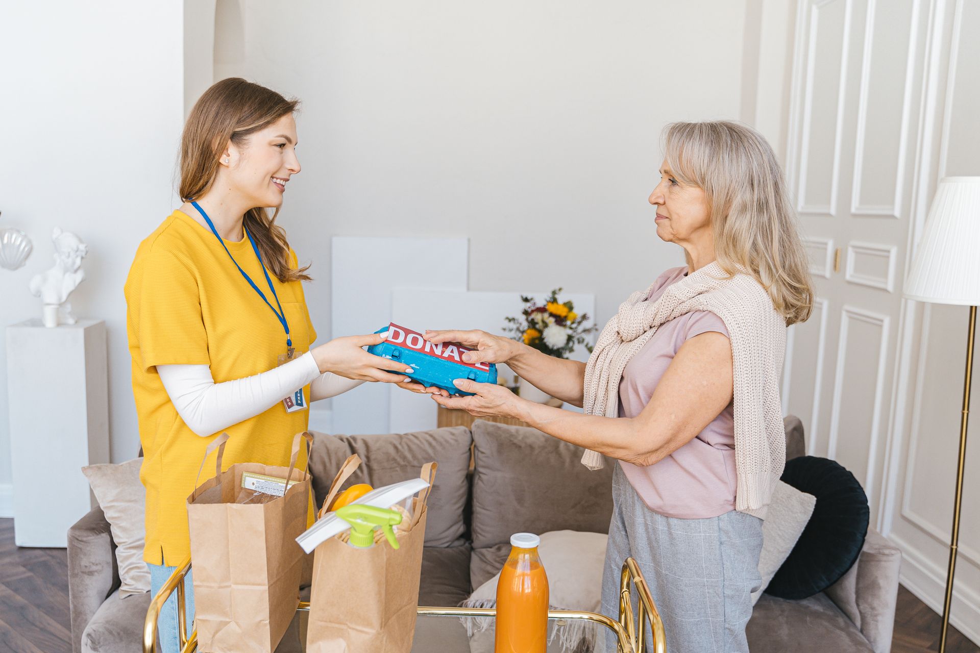 A young volunteer hands a donate box to a smiling senior woman in a warm living room, both standing face to face.