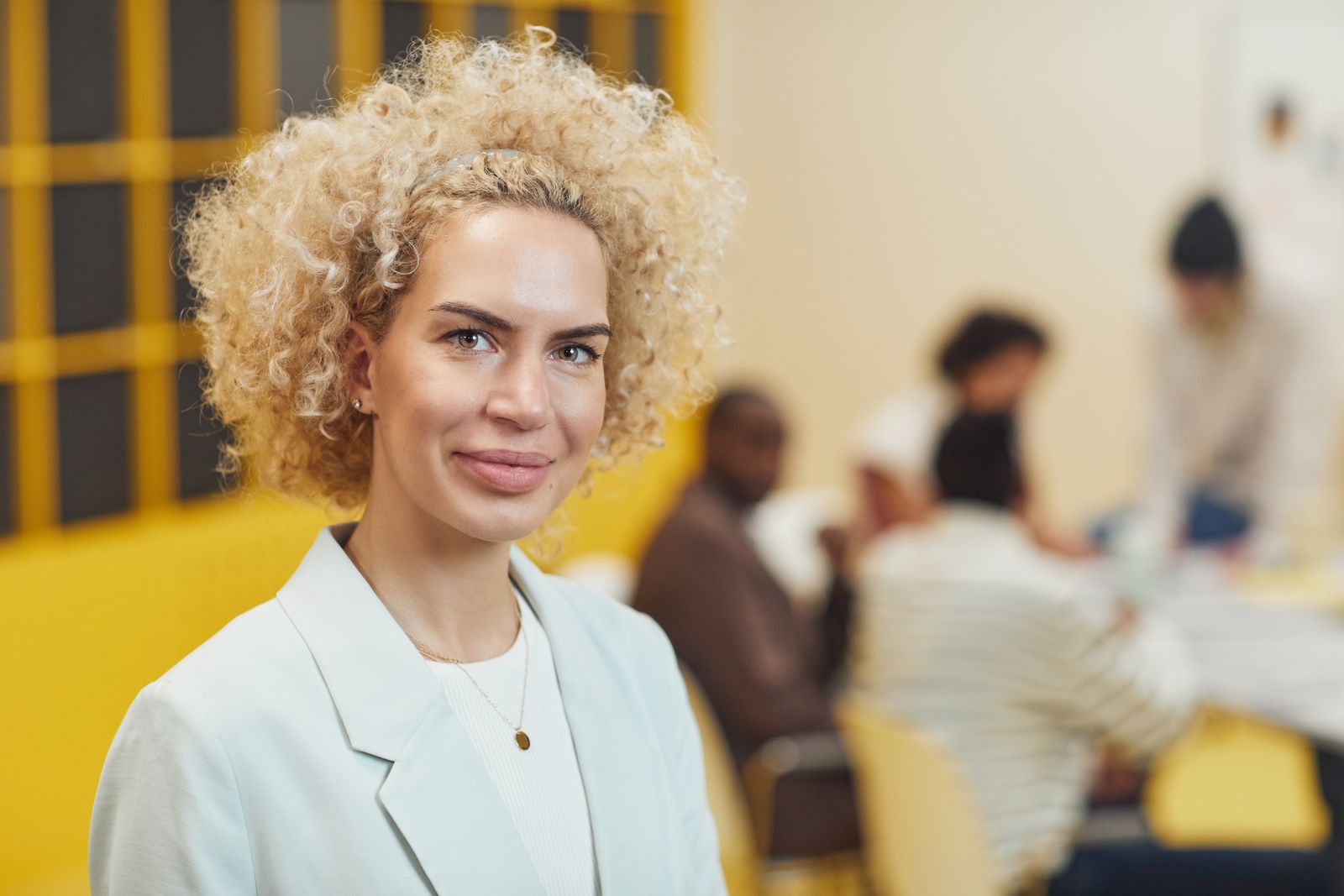 A friendly mixed-race woman with a blonde afro in a light blazer smiling at the camera, with a softly blurred team meeting in the background, used as the Volunteer Coordinator portrait.