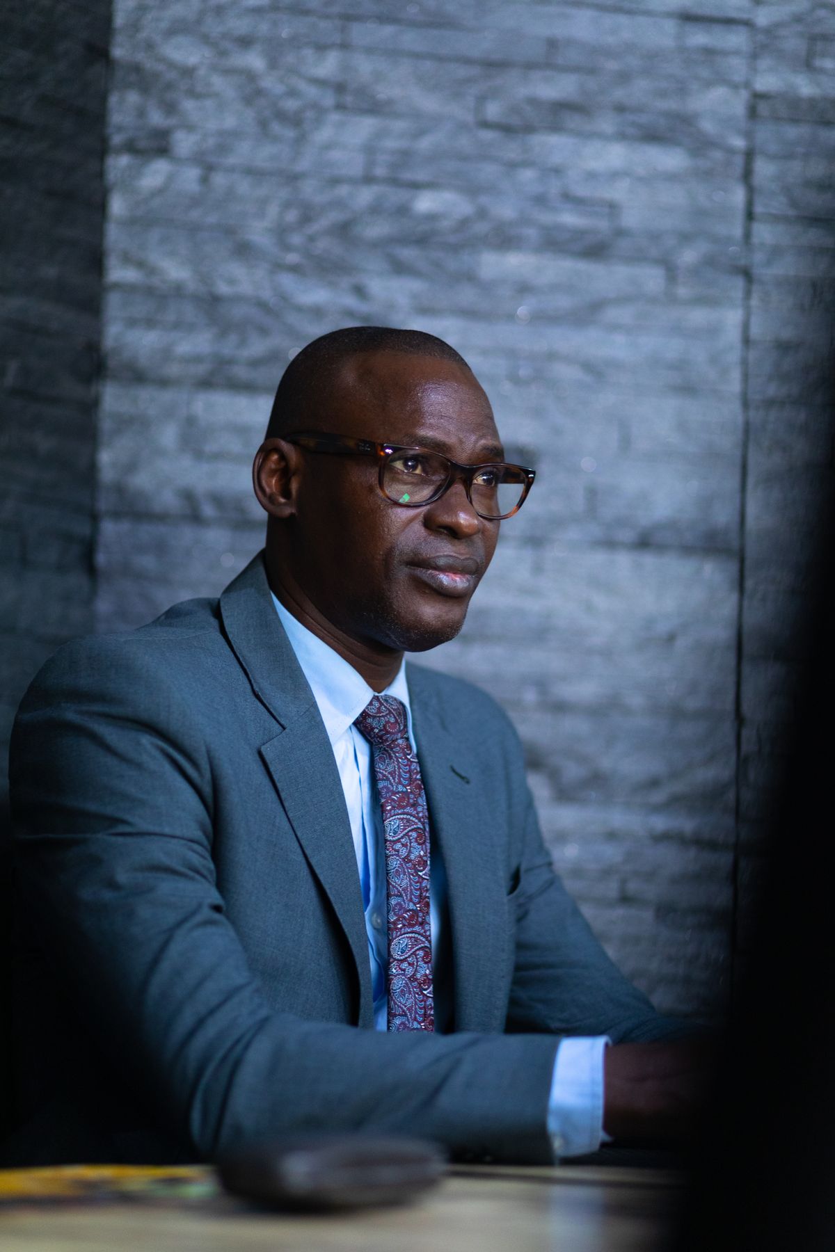 A professional Black man in a gray suit and glasses, looking composed and dedicated against a textured stone wall, used as the Program Director portrait.