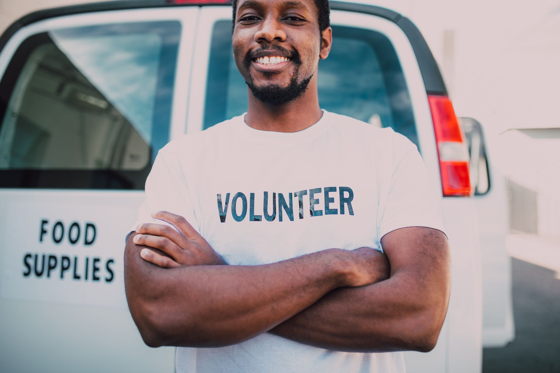 A confident male volunteer in a white volunteer t-shirt standing with arms crossed in front of a food supplies van, smiling.