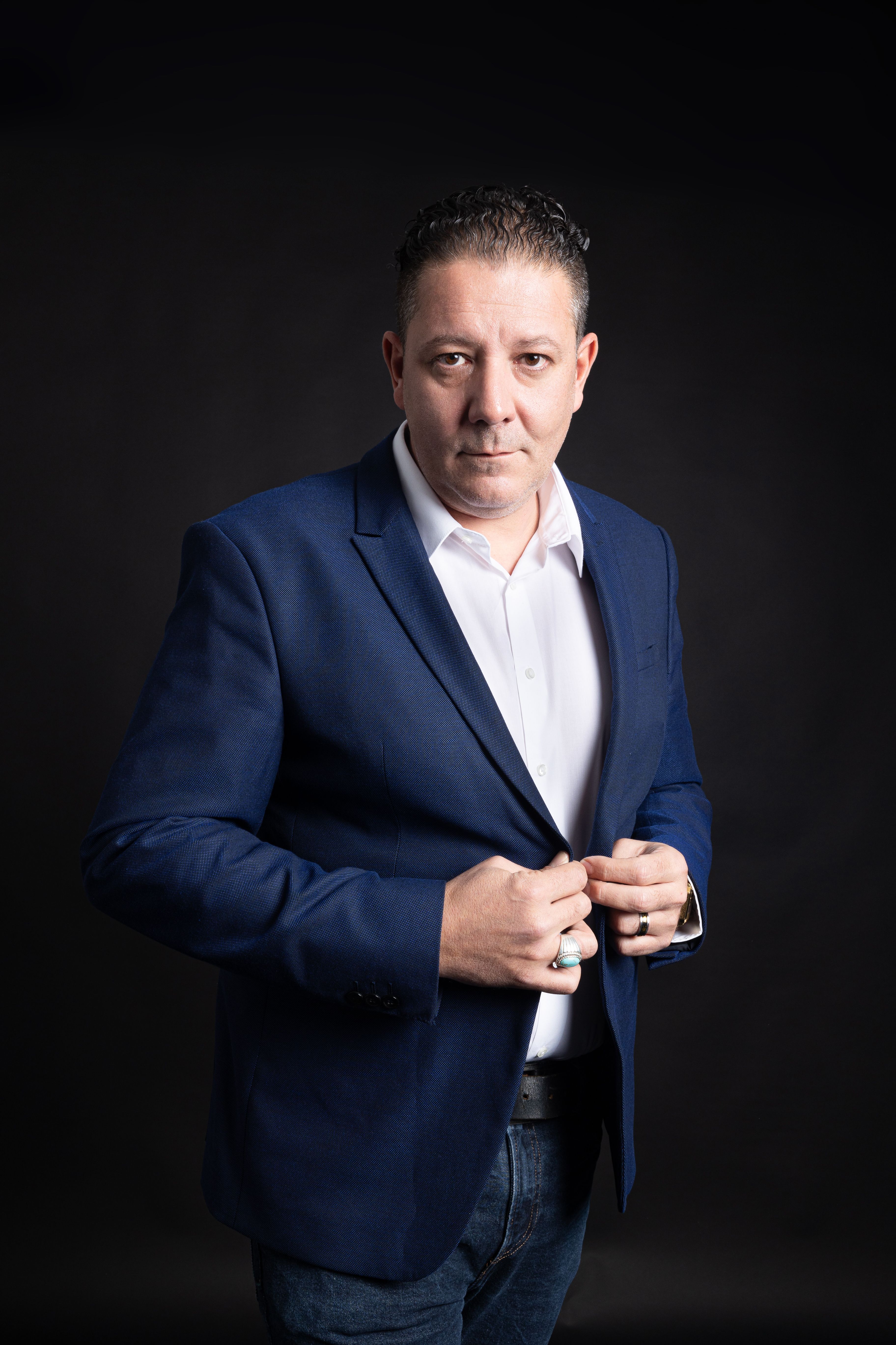 A confident middle-aged man in a navy blazer and white shirt against a dark studio background.