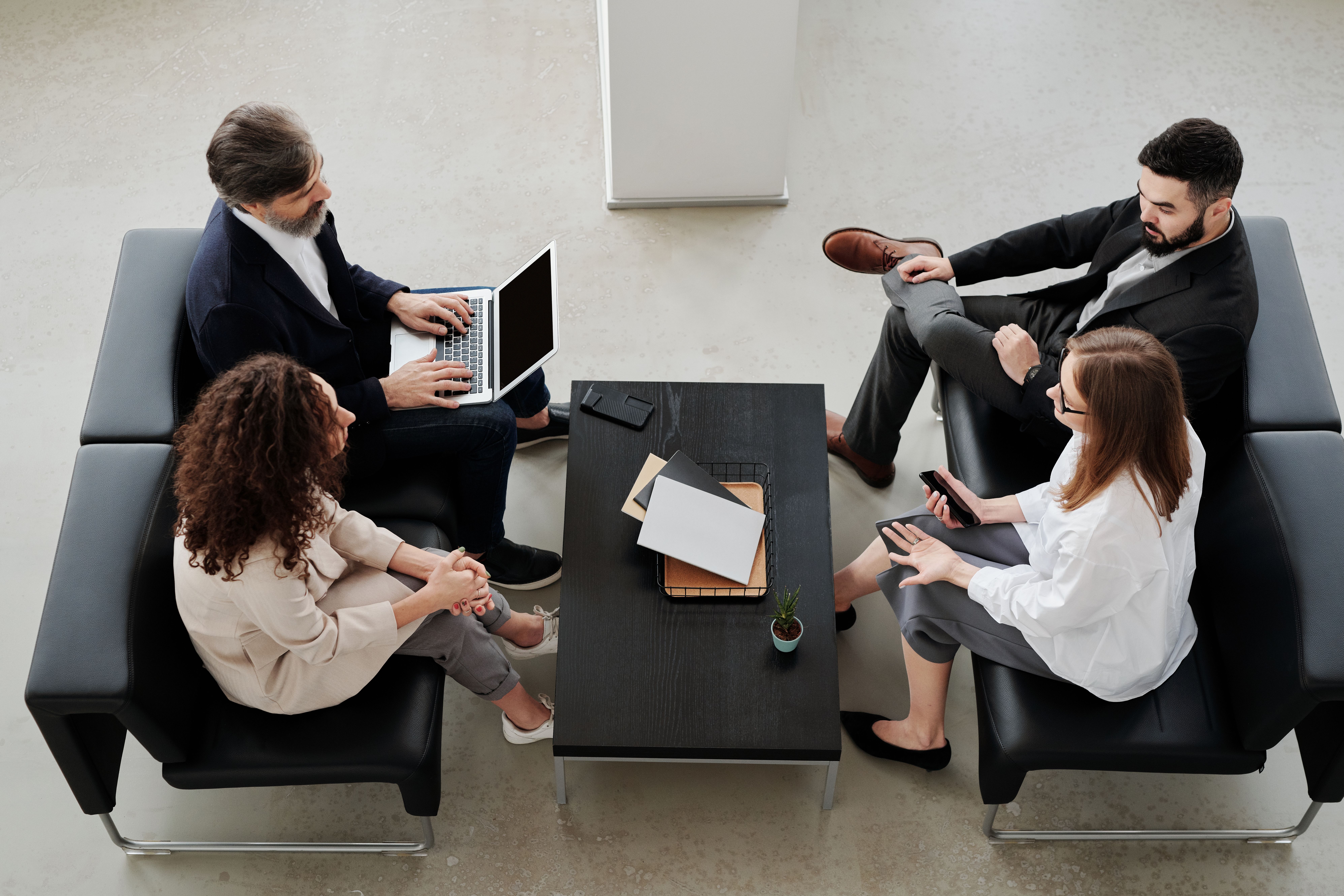 An overhead view of four professionals meeting on black leather sofas around a dark wood coffee table in a modern office.