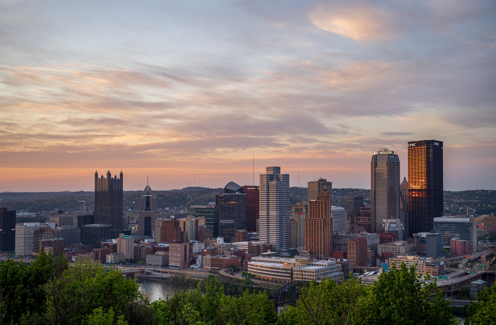 An aerial view of a mid-size American city downtown skyline at sunset, with golden light on the buildings and green trees in the foreground.