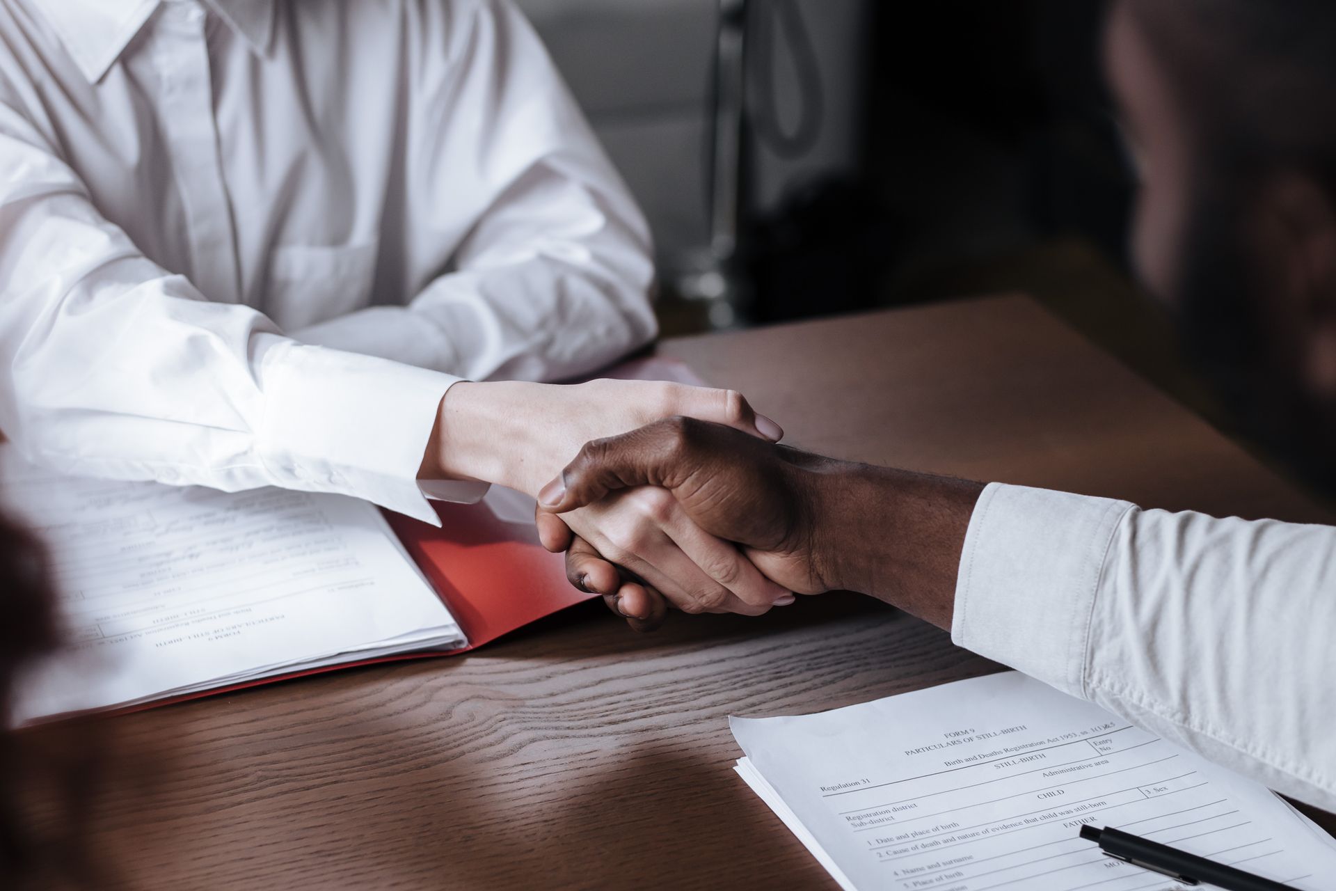 A close-up of two professionals of different ethnicities shaking hands over a printed contract on a wooden table.