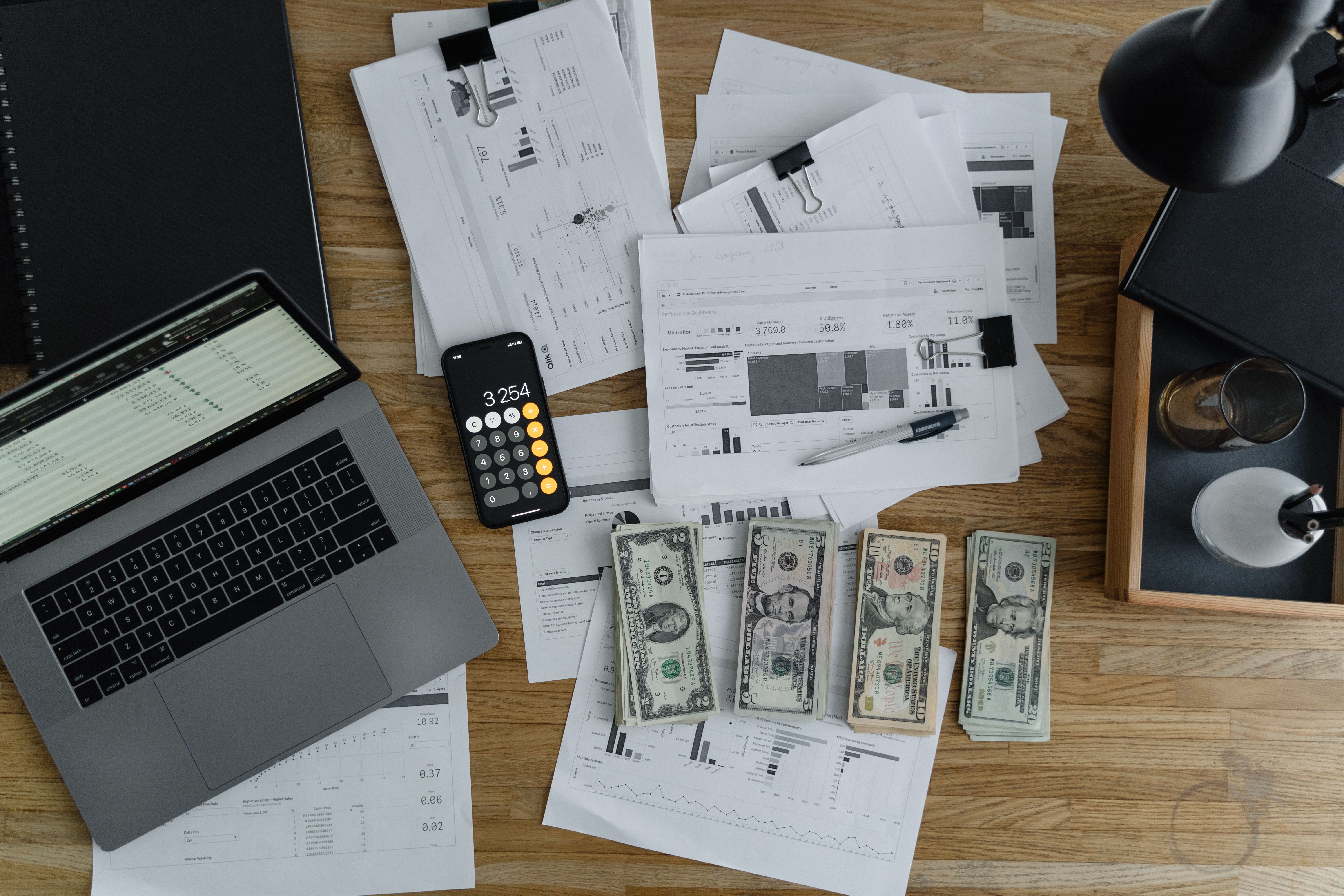 An overhead view of a laptop, printed financial reports with charts, a calculator, and dollar bills on a wooden desk.