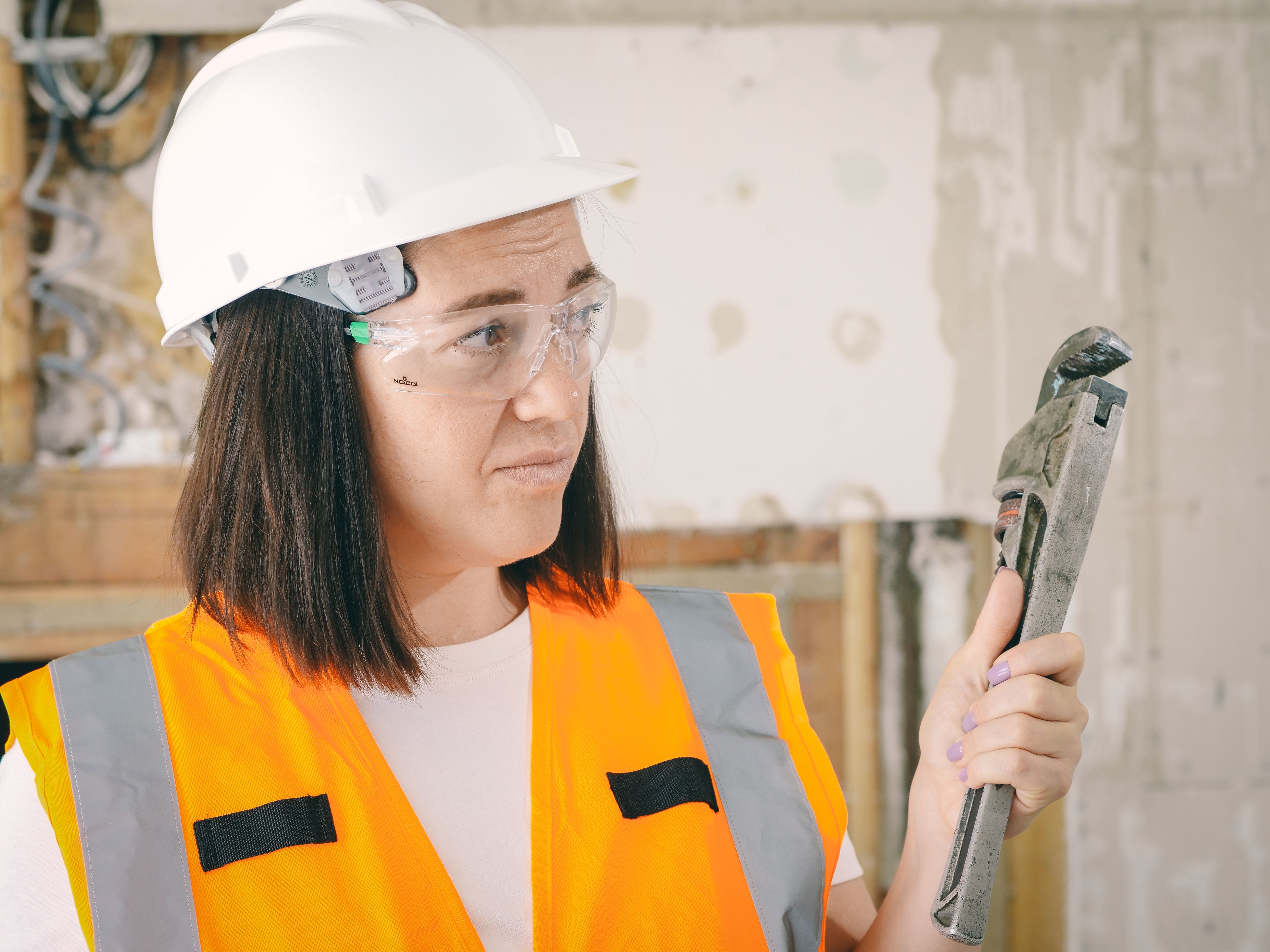 A maintenance professional in a hard hat, safety glasses, and high-visibility vest holding a pipe wrench during a residential repair.