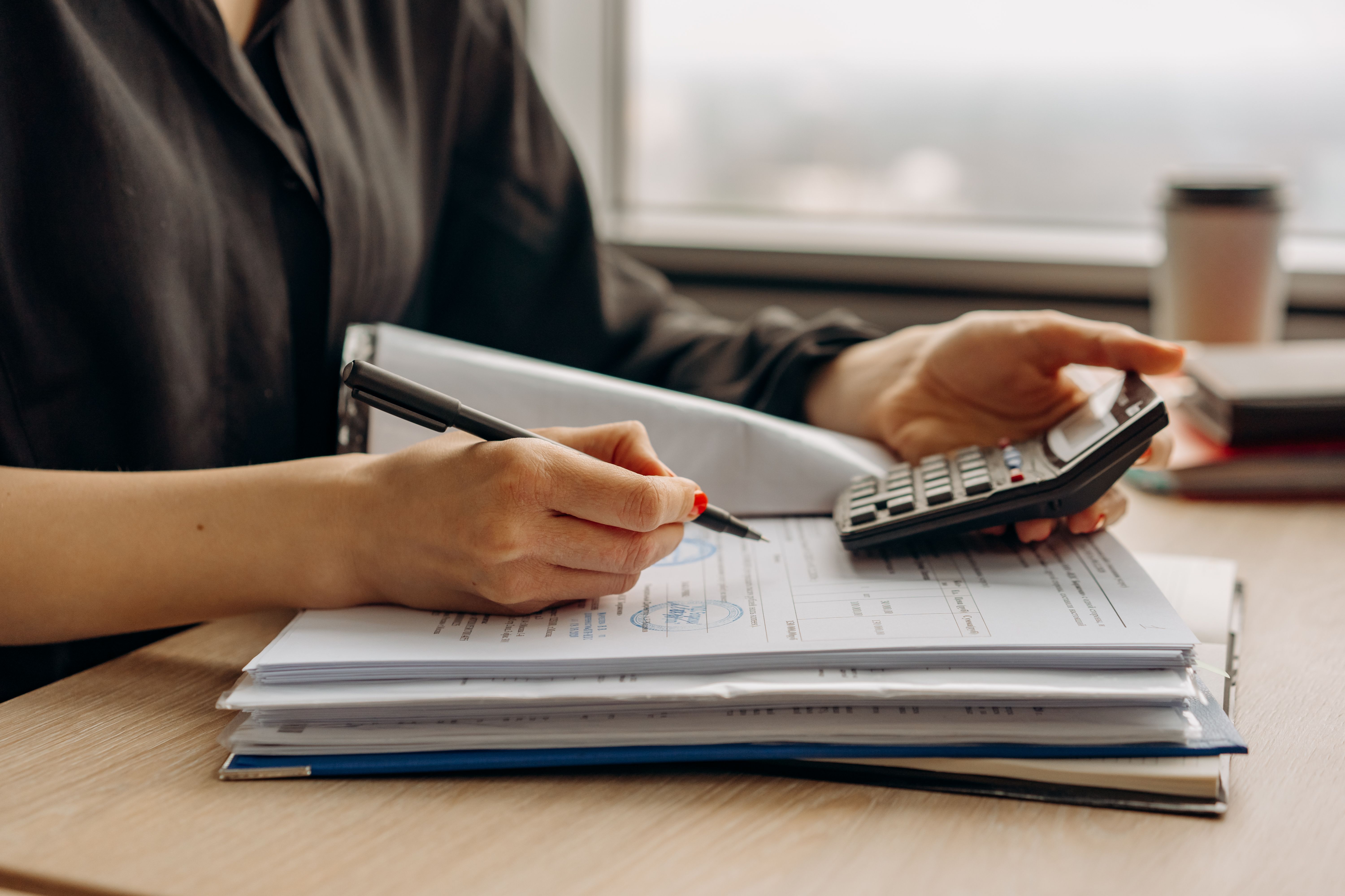 A person using a calculator and pen to review a stack of financial documents on a desk.