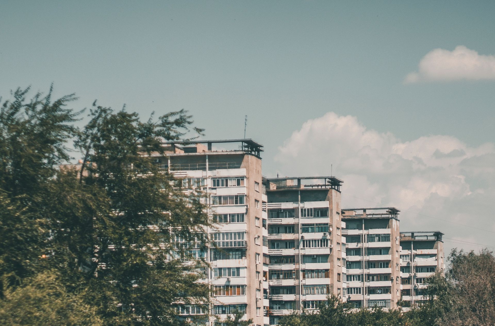 A row of mid-rise apartment buildings behind leafy green trees under a blue sky with light clouds.