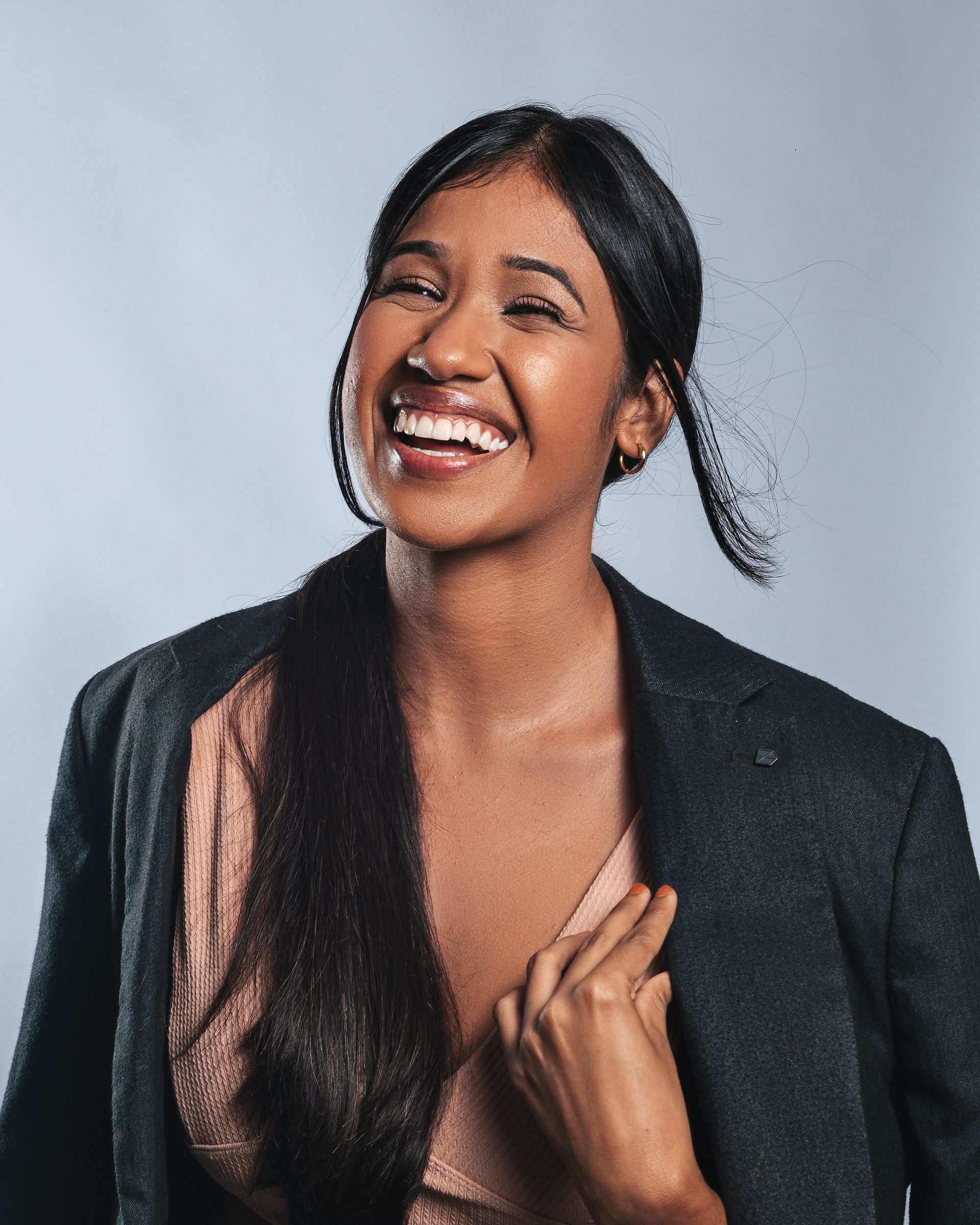 A joyful young woman in a dark blazer laughing with her hand on her chest, against a soft blue-gray background.