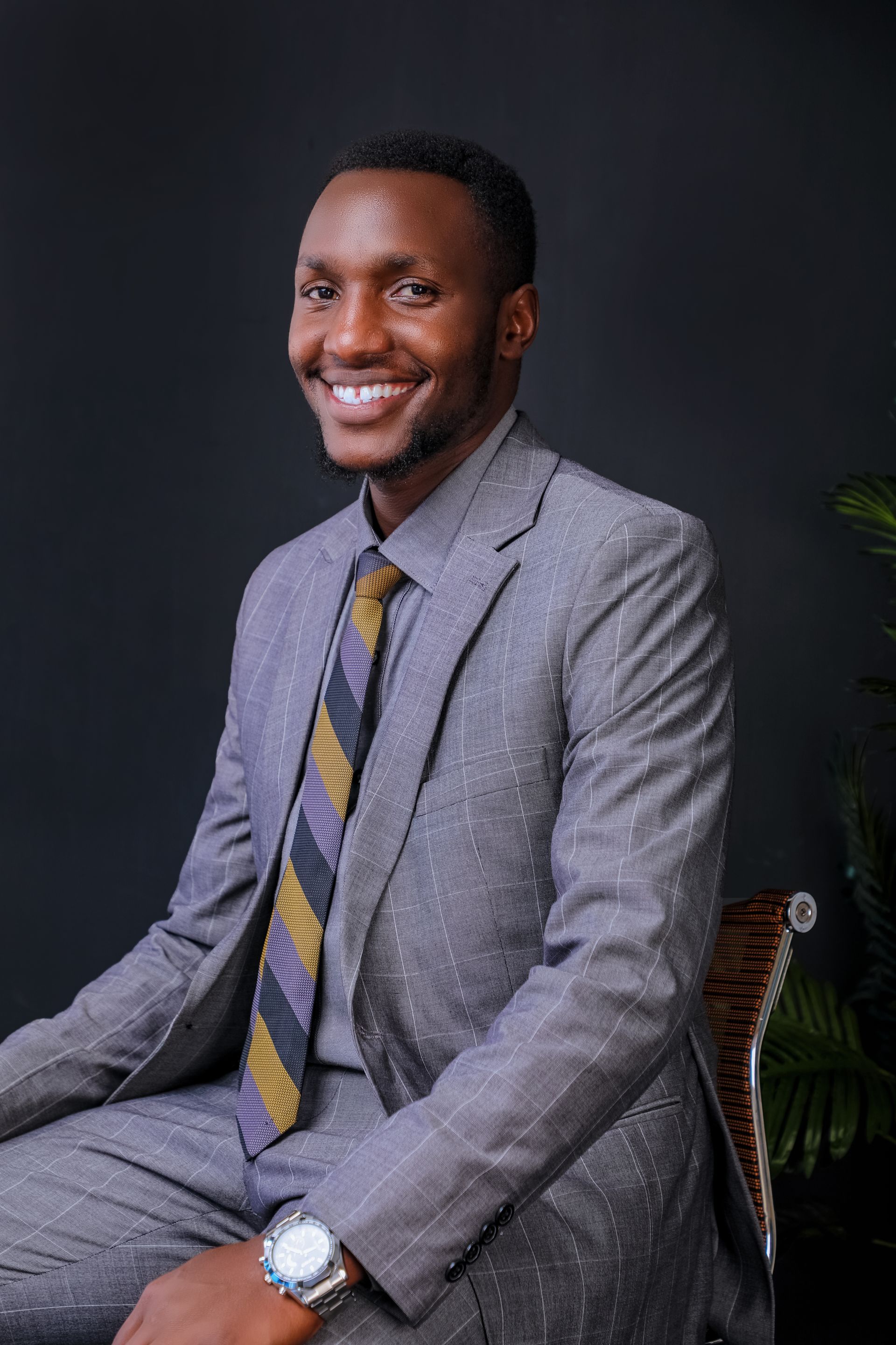 A smiling businessman in a gray suit and gold striped tie sitting in a chair against a dark studio background.