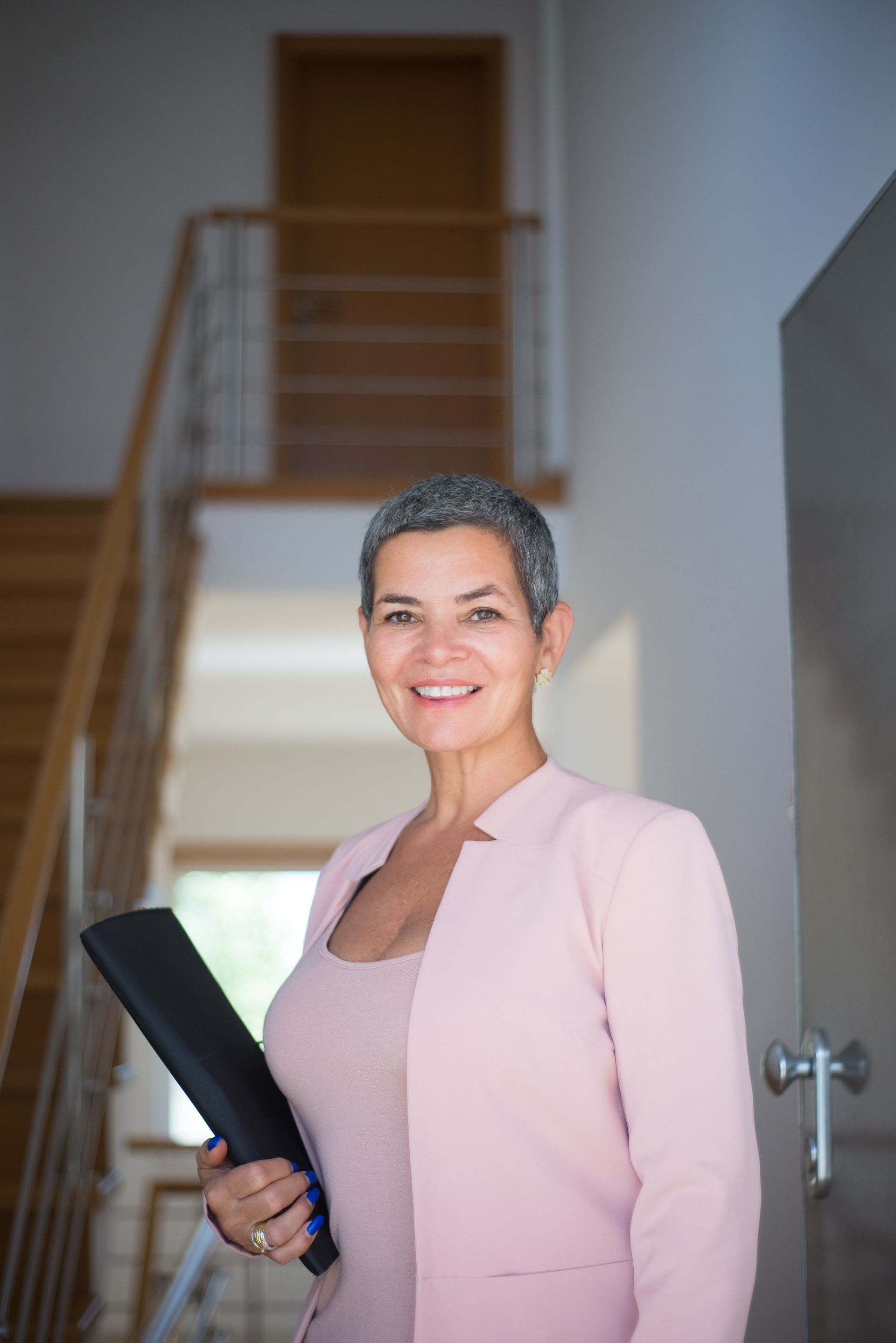 A smiling woman with short silver hair in a pink blazer holding a folder in a modern home with a wooden staircase behind her.