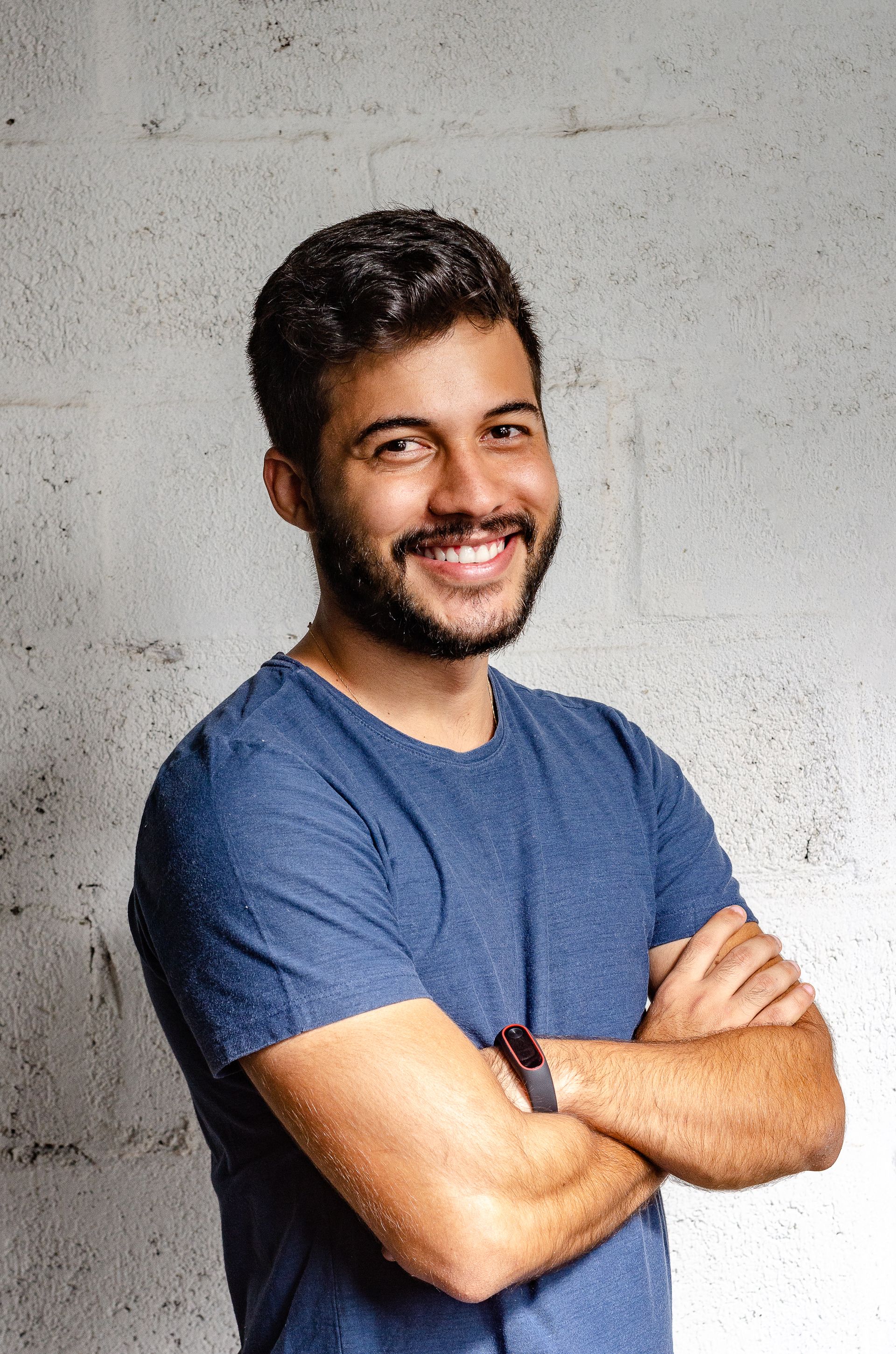 A smiling young man with a beard in a blue t-shirt, arms crossed, standing in front of a gray stone wall.