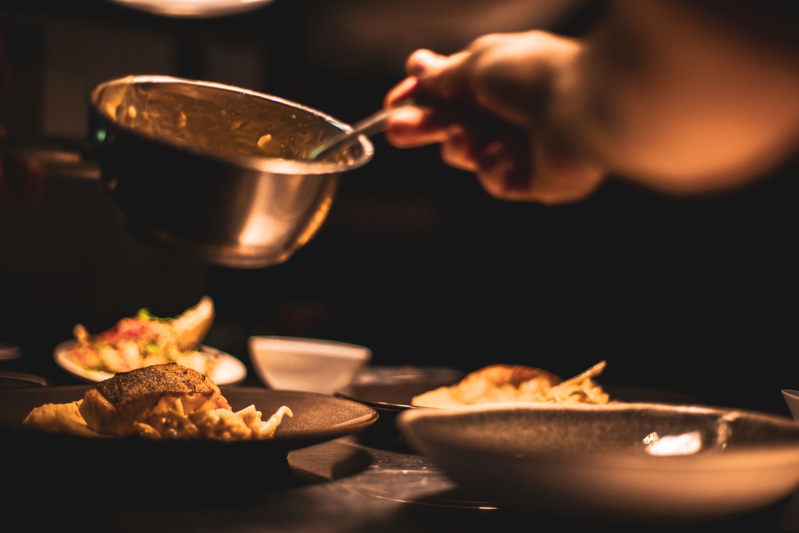 A chef pours sauce from a copper saucepan over plated dishes on a dark kitchen pass in warm amber light.