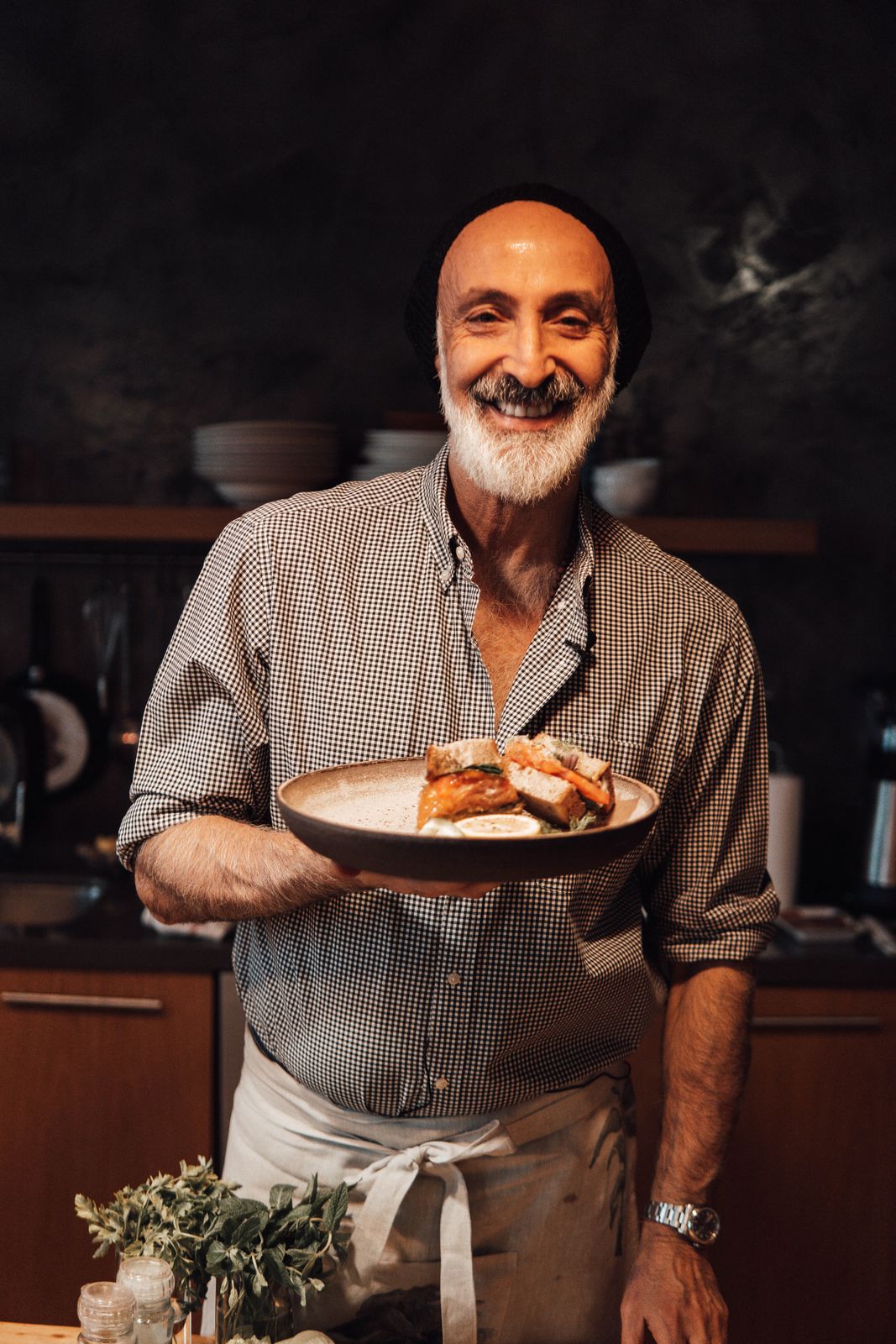 A confident and friendly older male chef with a white beard smiles warmly while holding a plated sandwich in a dim restaurant kitchen.