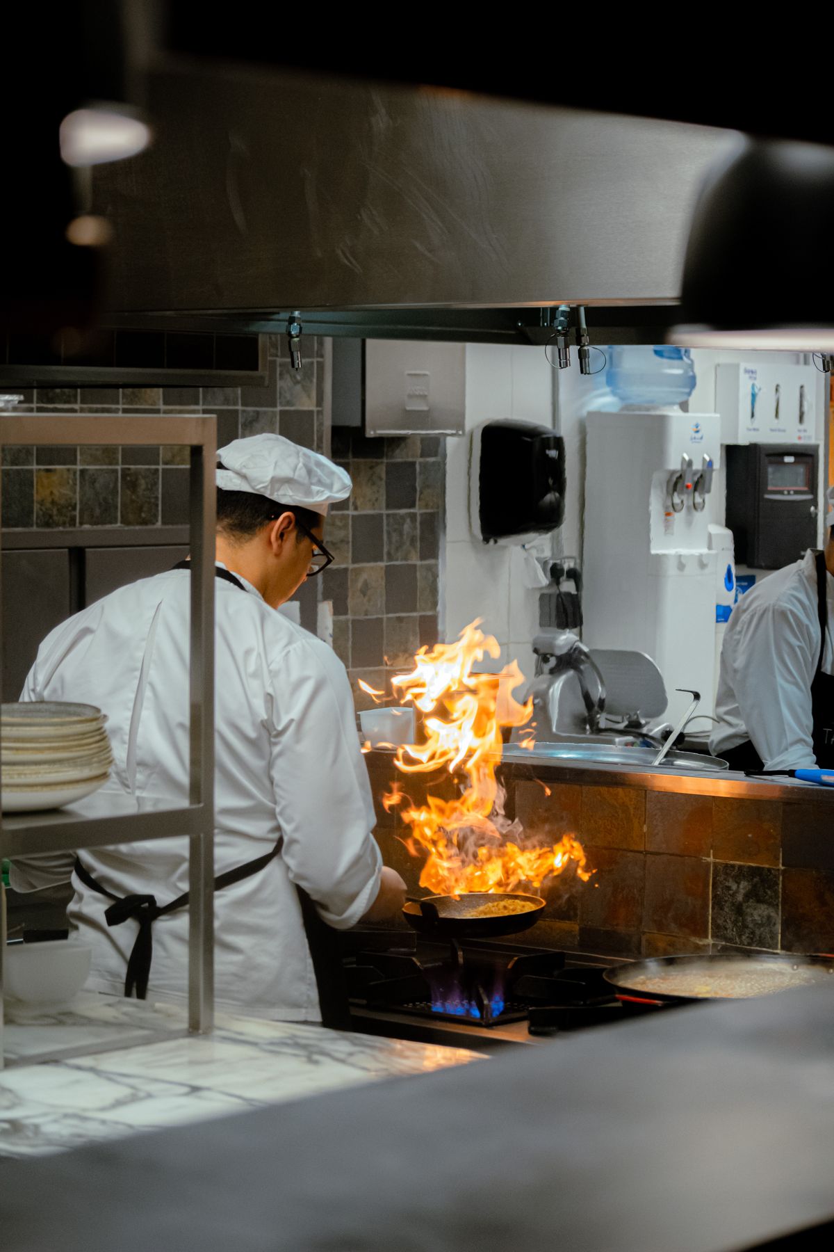 A chef in white coat and hat cooking on the line, with flames leaping from a saute pan at The Steel Hearth.