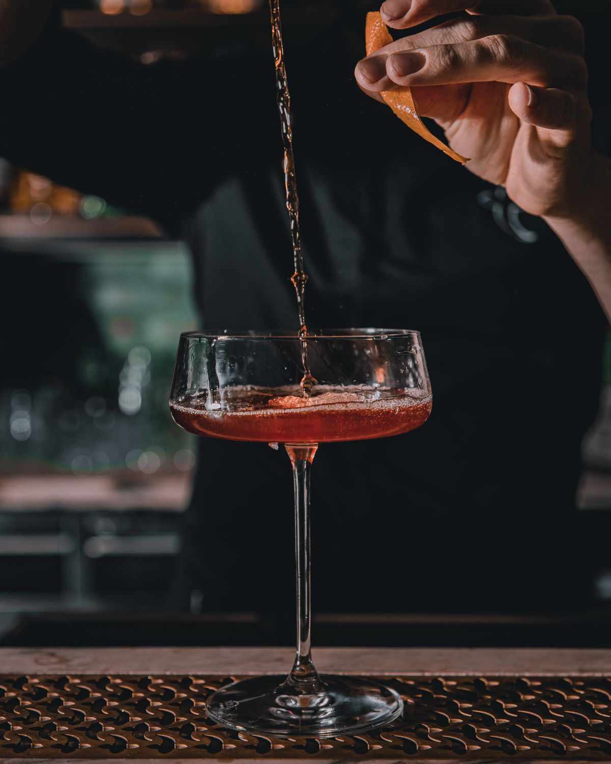 A bartender pouring a craft cocktail into a coupe glass, with an orange-peel twist held above, at The Steel Hearth.
