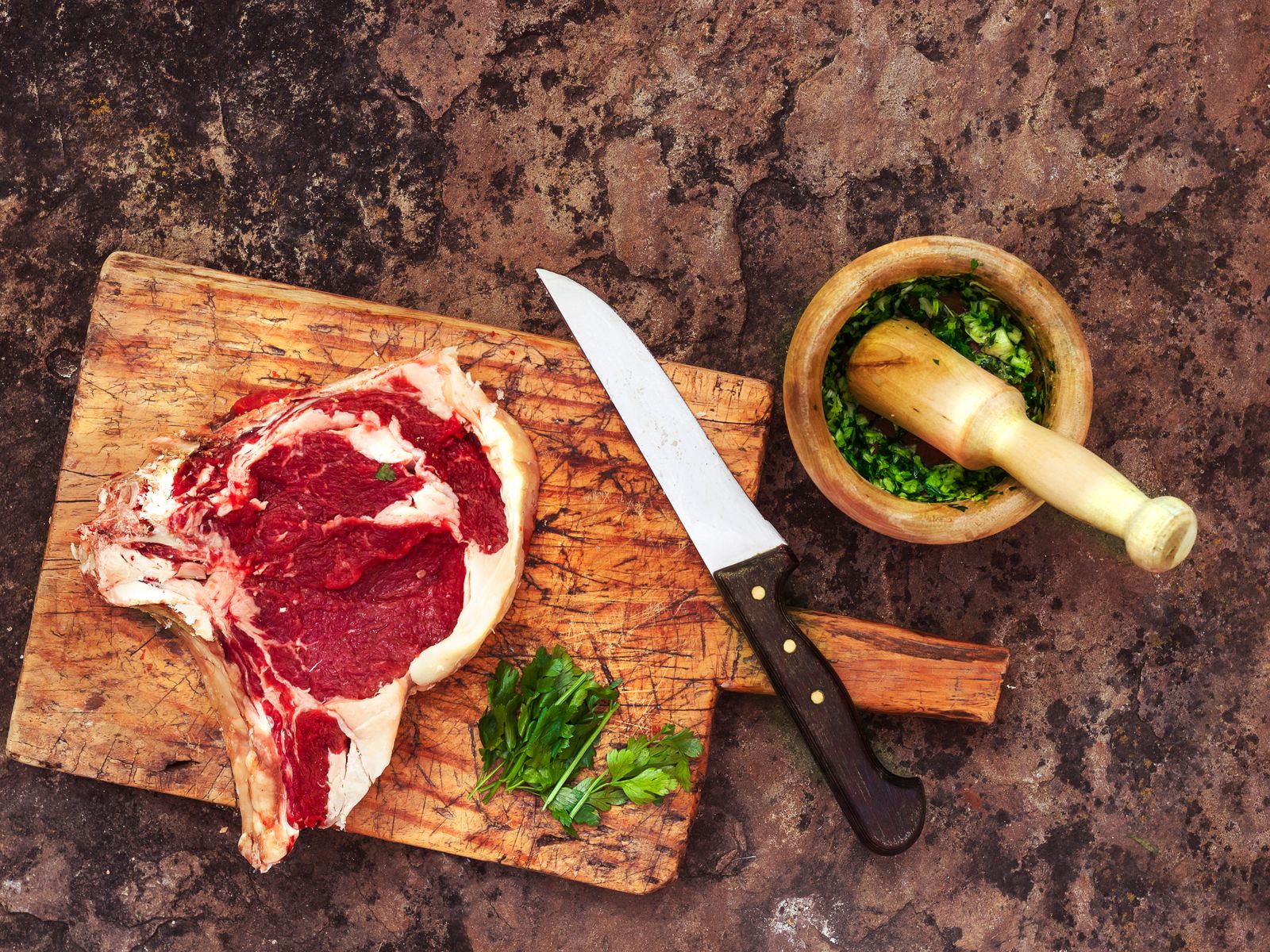 A raw T-bone steak on a wooden cutting board with a chef's knife, fresh parsley, and a mortar and pestle of chopped herbs, laid out on a dark prep counter.