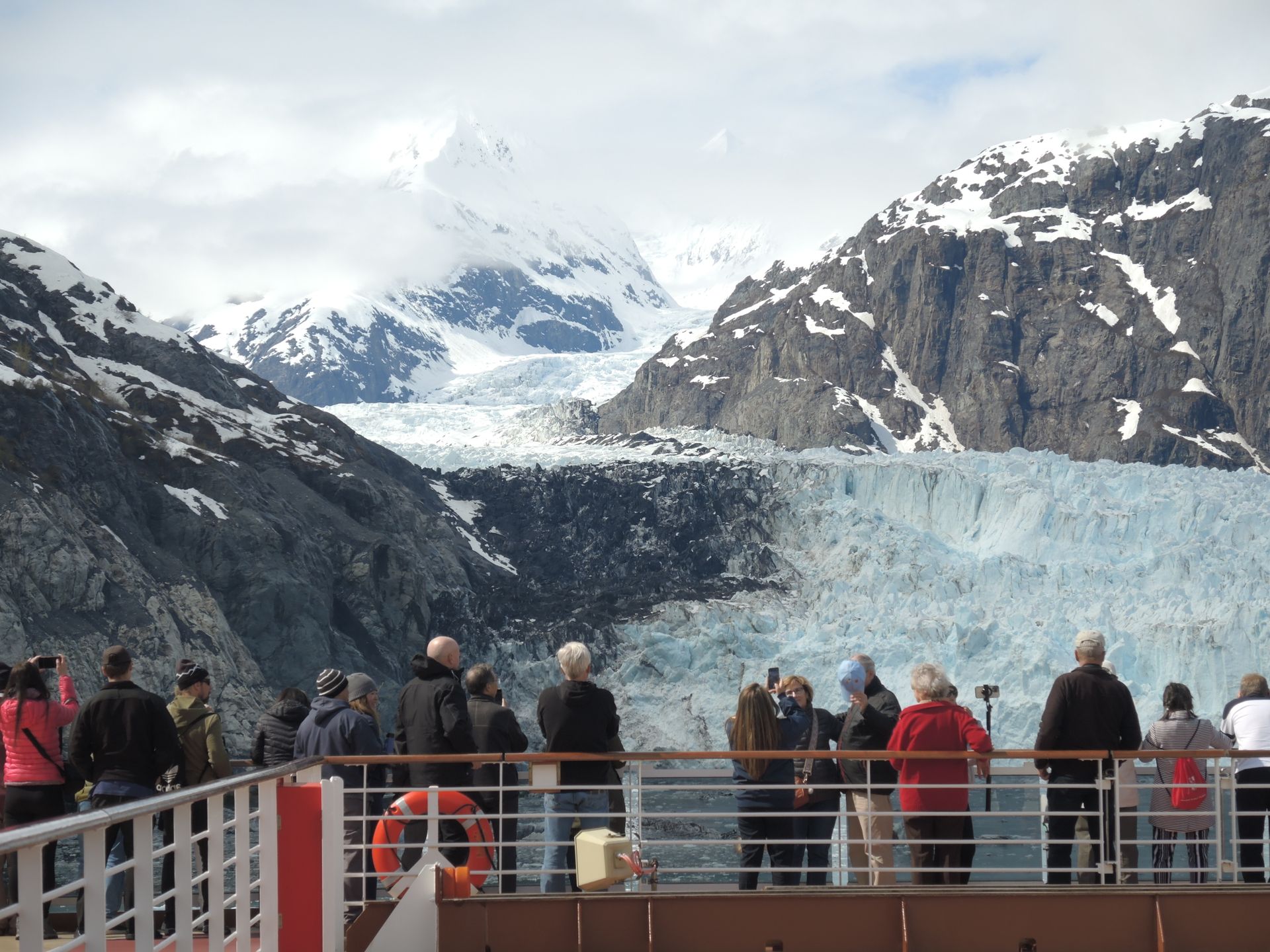 A group of tourists on a cruise ship deck photographing a massive blue tidewater glacier surrounded by snow-capped mountains in Alaska.
