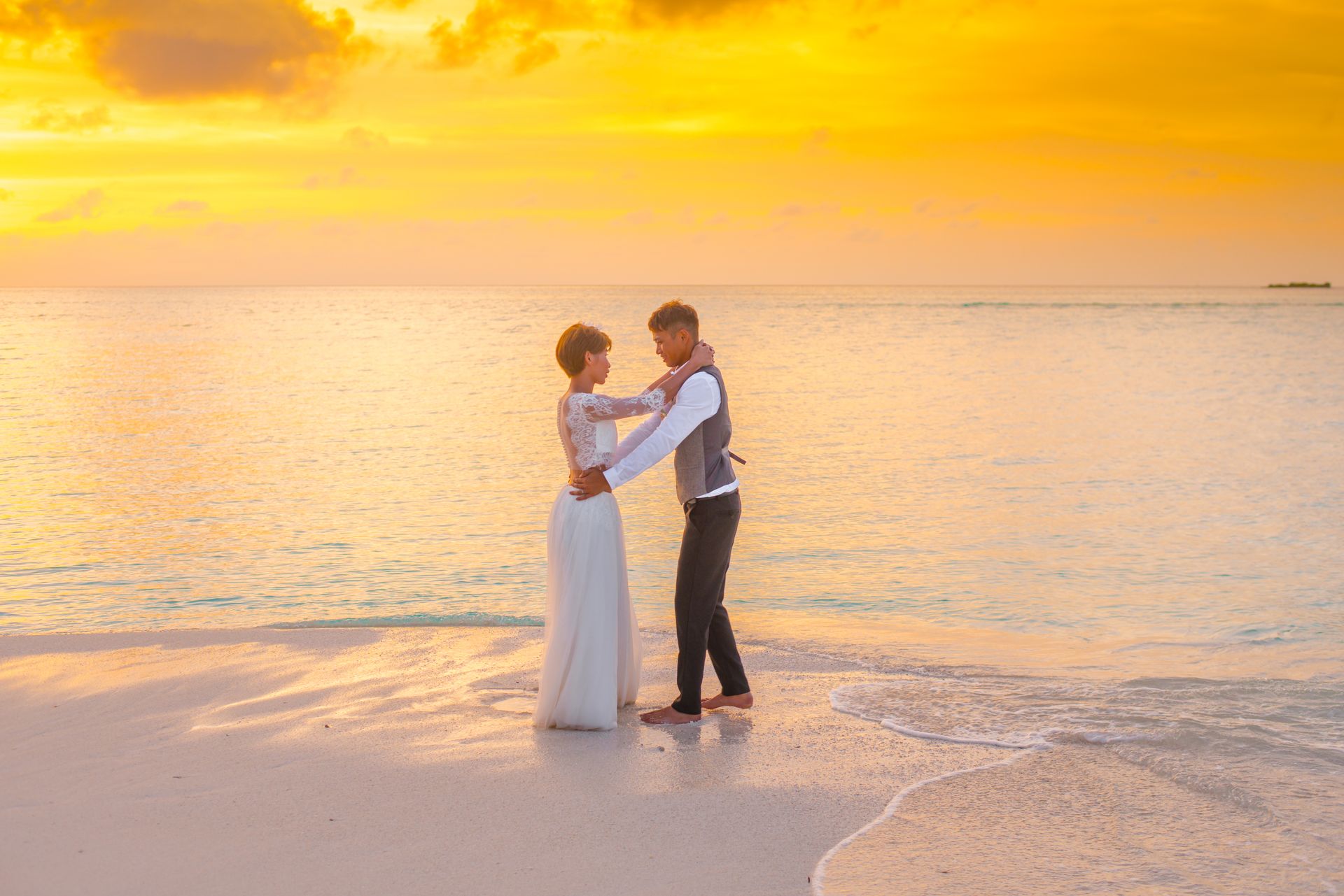 A bride in a white lace gown and groom in a gray vest embrace on a tropical beach at golden hour, with the warm sunset sky reflected in the calm ocean behind them.