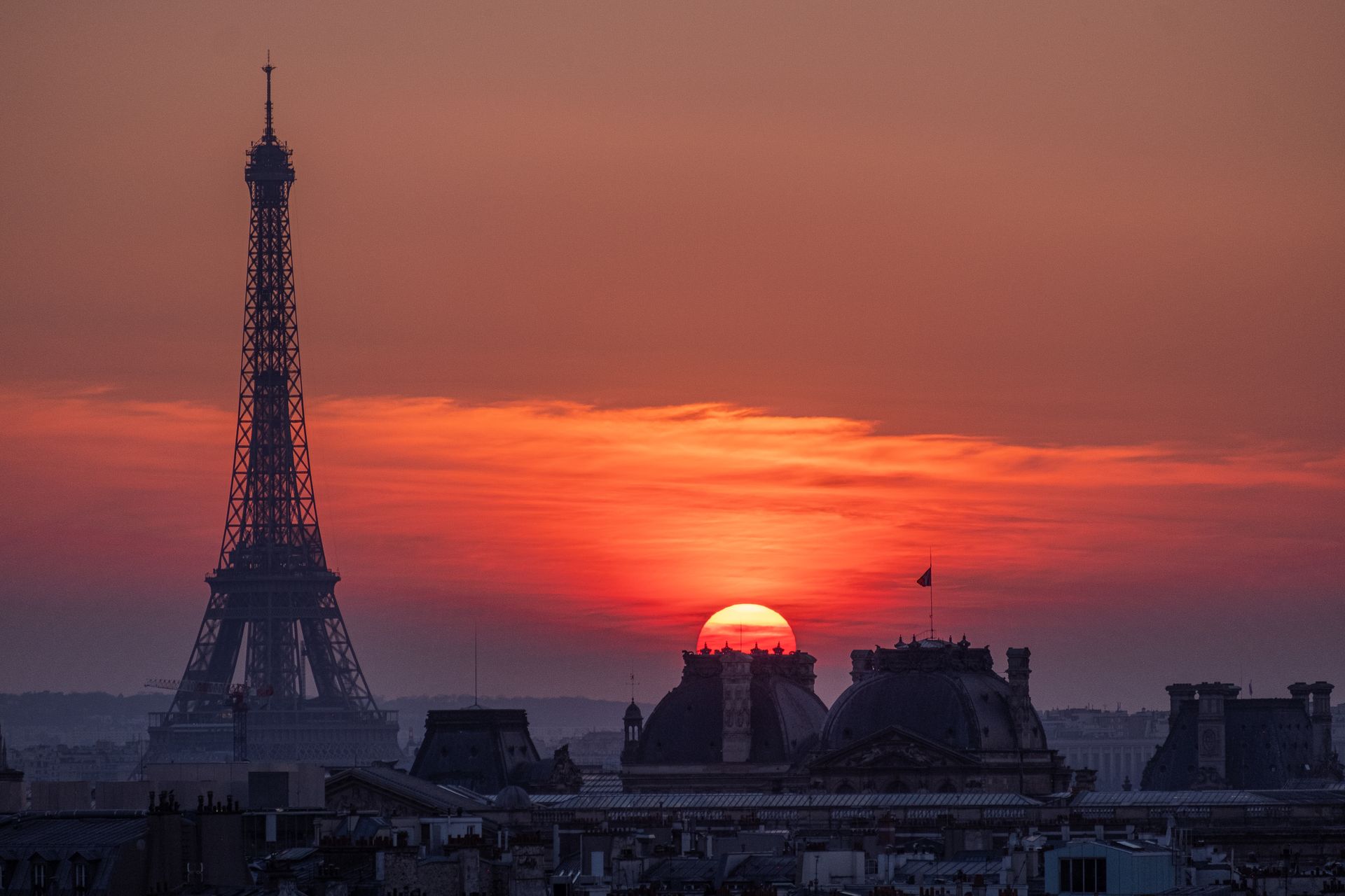 A silhouette of the Eiffel Tower at sunset in Paris, with a red sun setting behind the domes and rooftops of the city.