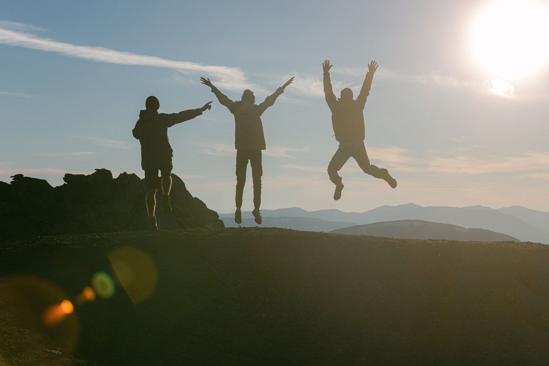 Three friends silhouetted against a sunset sky, captured mid-jump on a mountain summit with arms raised in celebration.