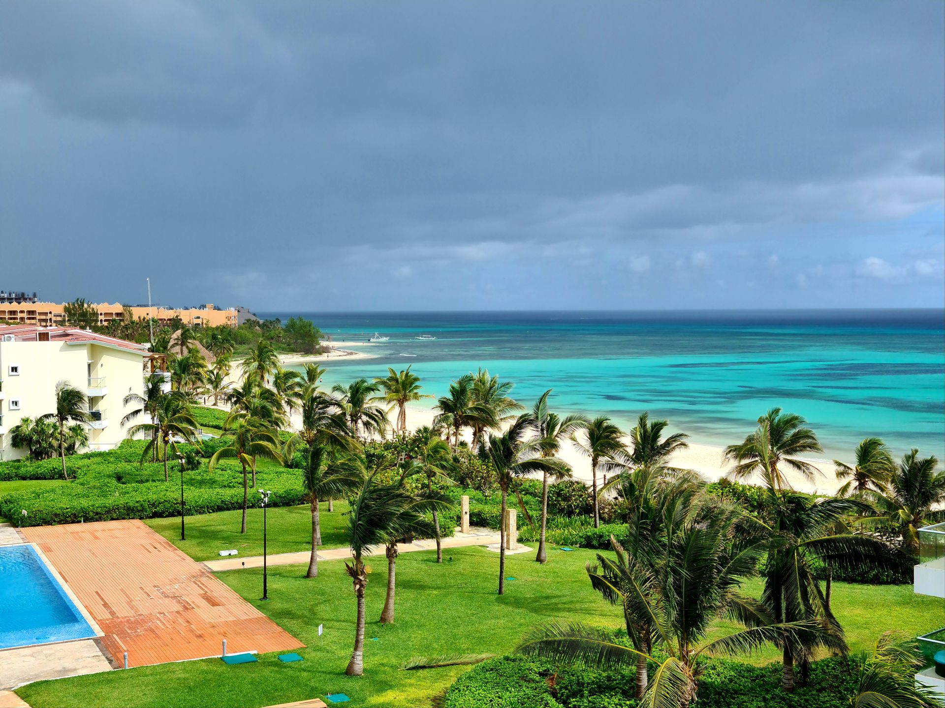 An aerial view of a beachfront resort with a pool, palm trees, white sand, and turquoise Caribbean water under a partly cloudy sky.