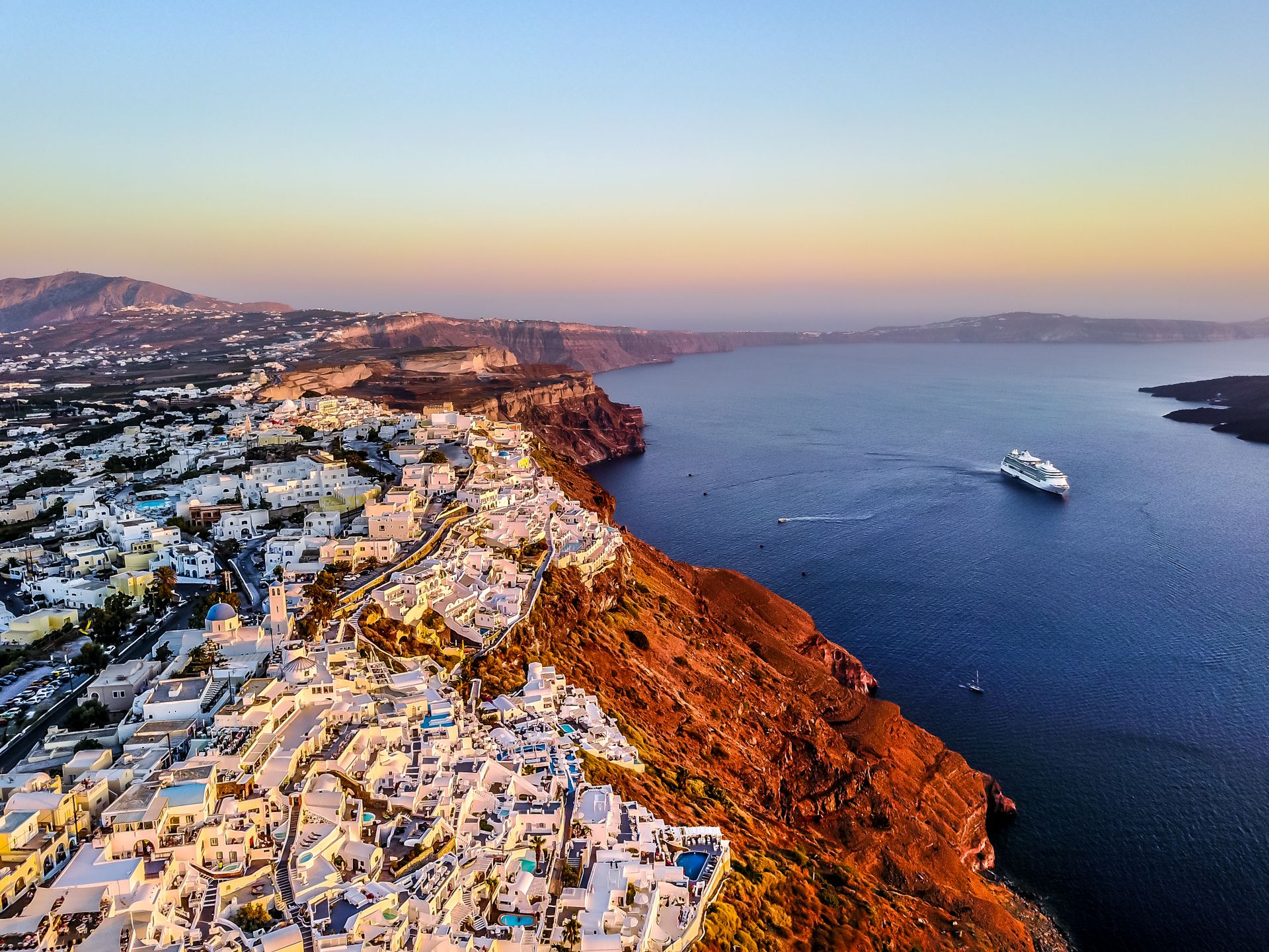 An aerial view of Santorini Greece at sunset, with white buildings and blue-domed churches on volcanic cliffs and a cruise ship sailing on the deep blue Aegean Sea.