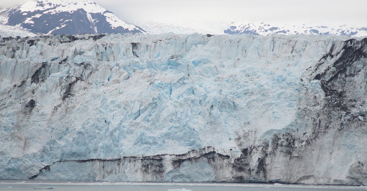 A massive blue Alaskan tidewater glacier face with snow-capped mountains rising behind and ice floating in the water below.