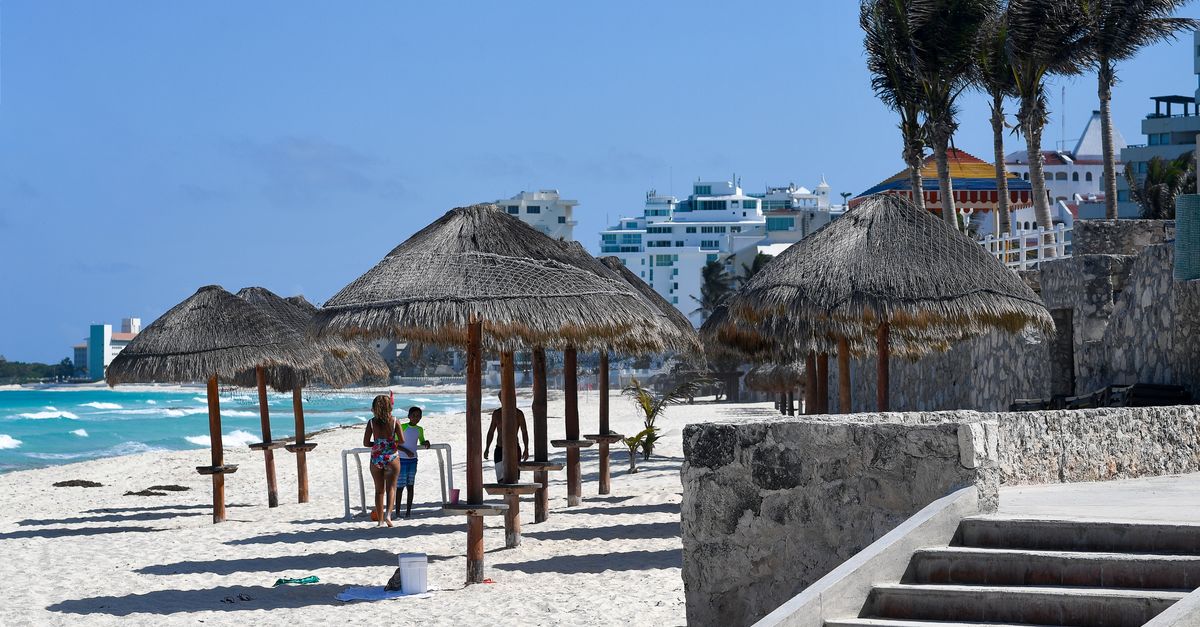 Thatched-roof palapa umbrellas on a white sand Cancun beach with turquoise Caribbean water and hotel buildings in the background.
