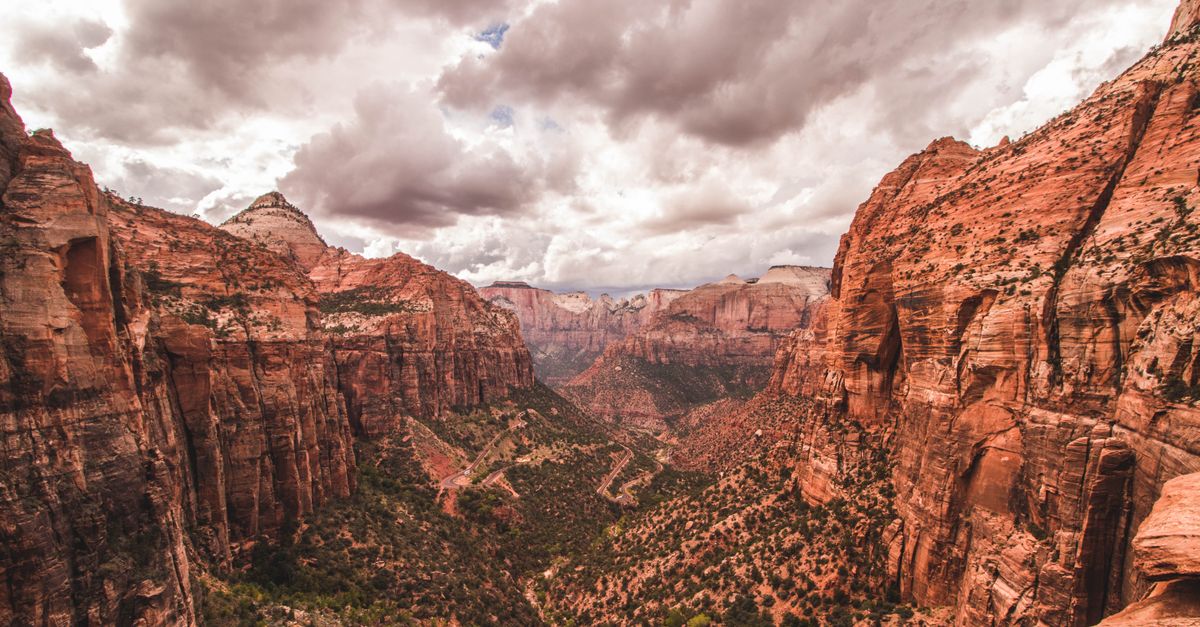 A dramatic red-rock canyon in Zion National Park, Utah, with towering sandstone cliffs and a switchback road winding down to the valley floor.