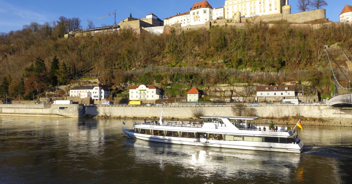 A white European river cruise boat on the Danube River in Passau, Germany, with a hilltop fortress and historic buildings rising above the riverbank.