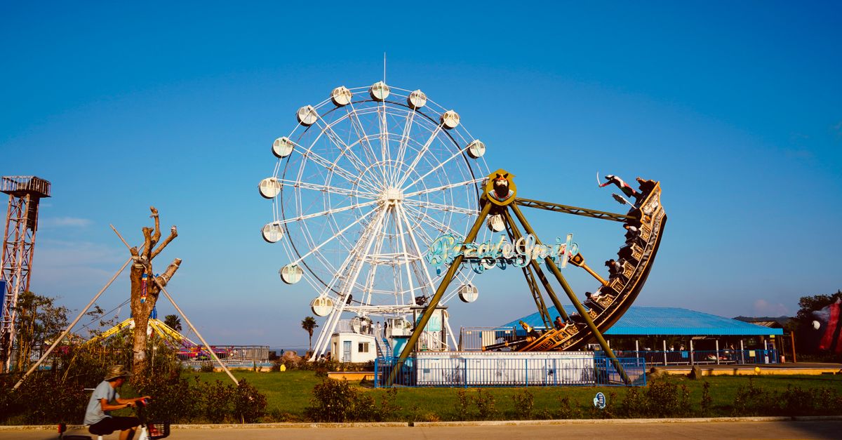 A colorful amusement park with a large white Ferris wheel and a pirate ship swing ride against a clear blue sky.