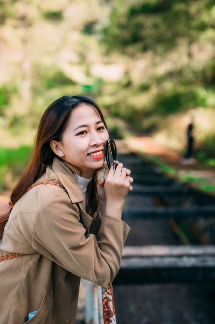 Sarah Ng, Travel Advisor at Steel City Travel, a warm East Asian woman in her 30s wearing a tan trench coat and holding sunglasses, smiling brightly in front of sunlit railway tracks.