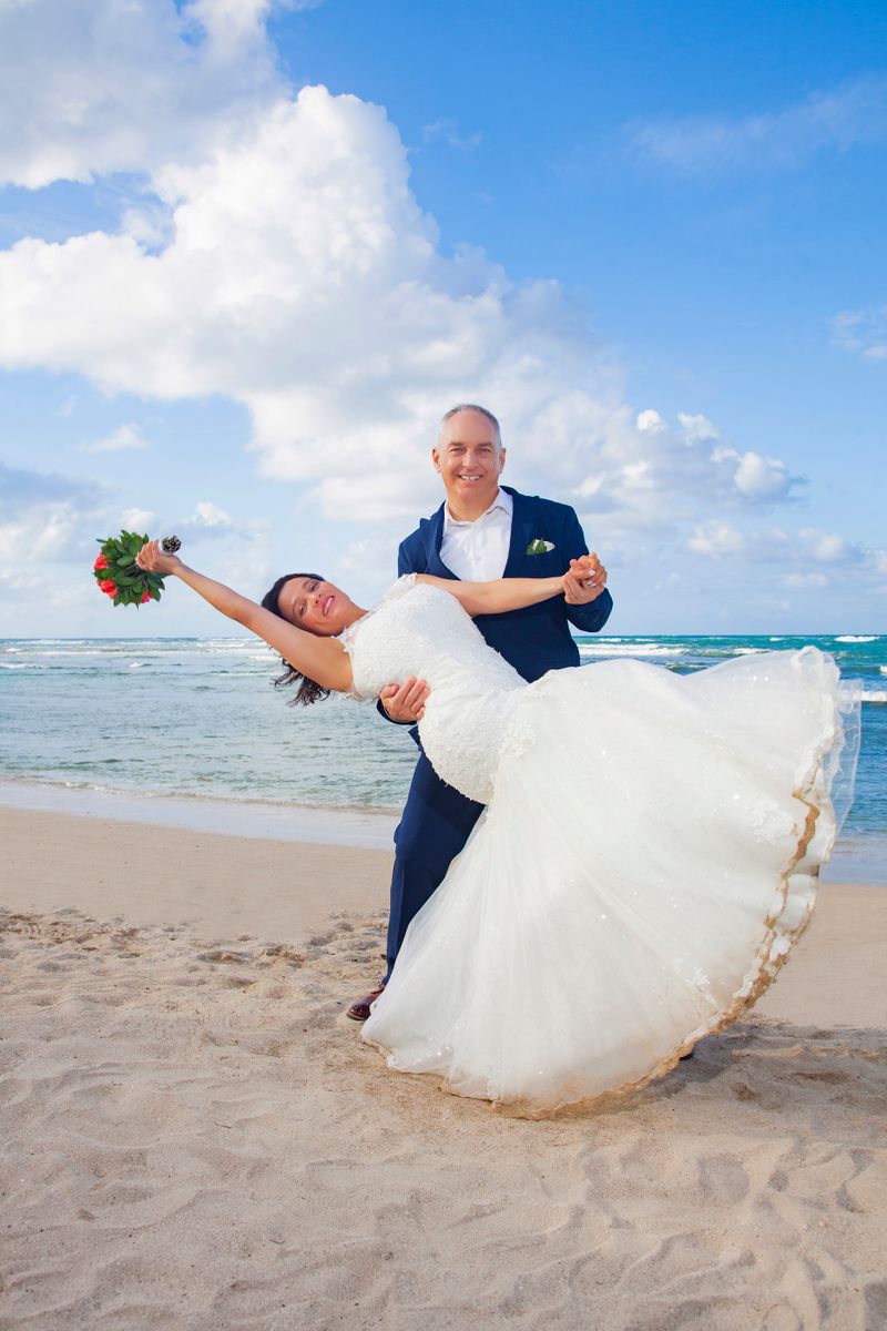 Jennifer and David K. on their destination wedding day in Jamaica, a joyful white couple in their 30s, the groom in a navy suit dipping the bride in a white lace gown on a Caribbean beach.