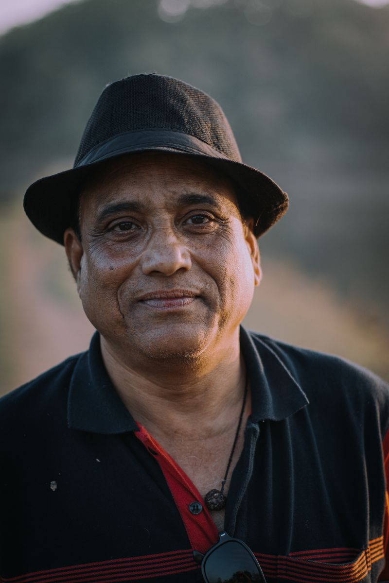 Michael T. after his Caribbean cruise, a warm and content South Asian man in his 50s wearing a black fedora and black polo with red trim, smiling in golden-hour light.