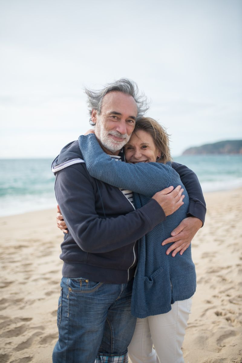 Robert and Linda M. after their 10-day Rome and Italy trip, a happy white couple in their 50s-60s embracing on a beach, the man with a gray beard and the woman in a sun hat.