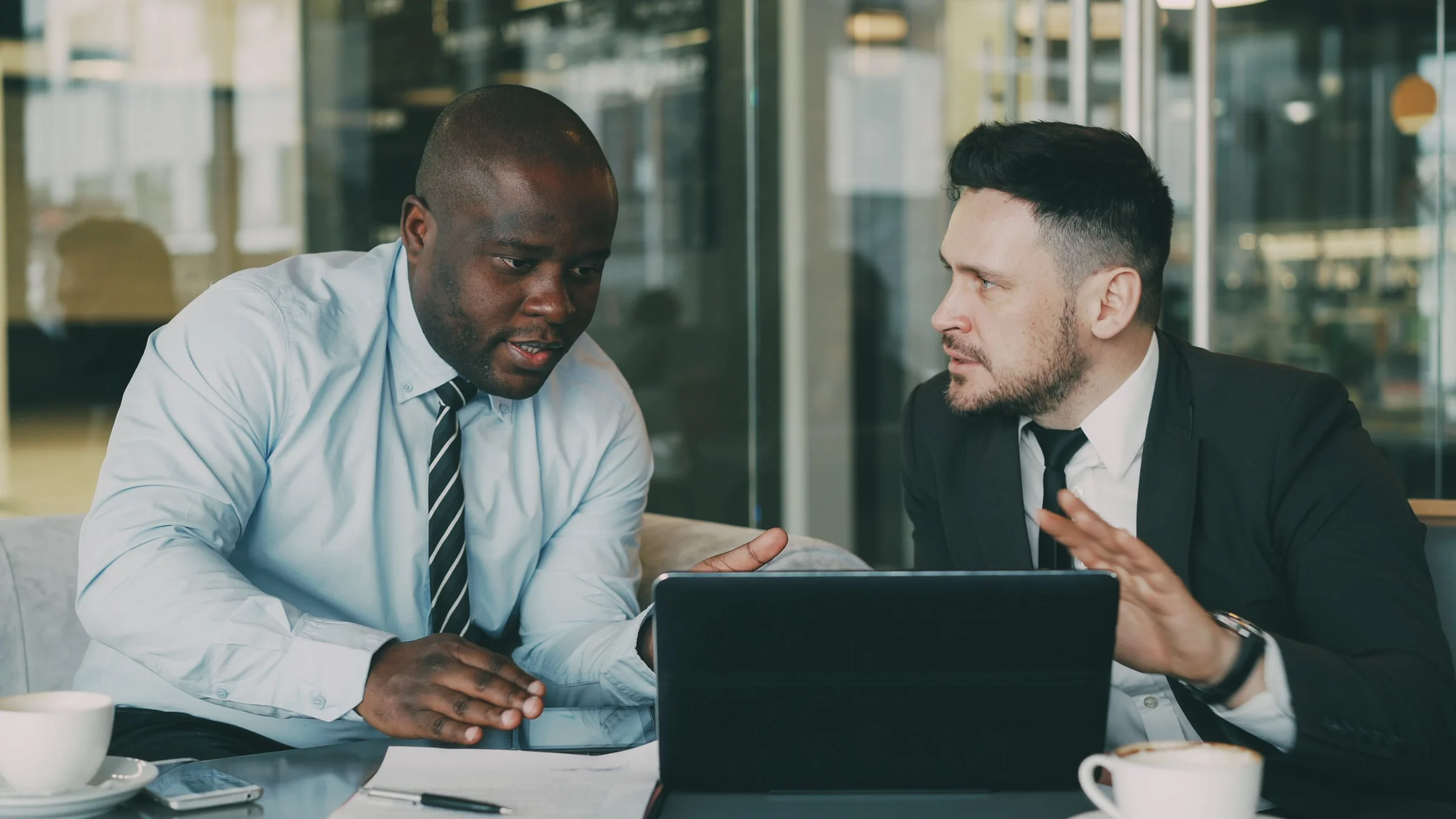 Two businessmen in smart formal attire discussing a project over a laptop in a glass-walled office lounge, coffee cups and paperwork on the table, cool blue-toned light.