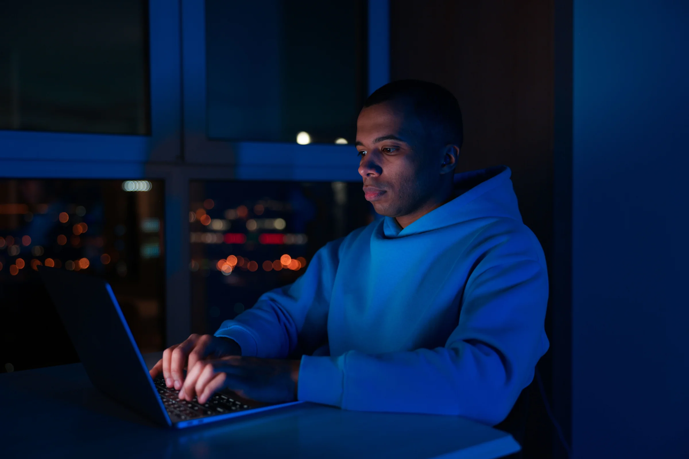 A programmer in a blue hoodie typing code on a laptop at night, deep blue light filling the room with warm city bokeh through the window.